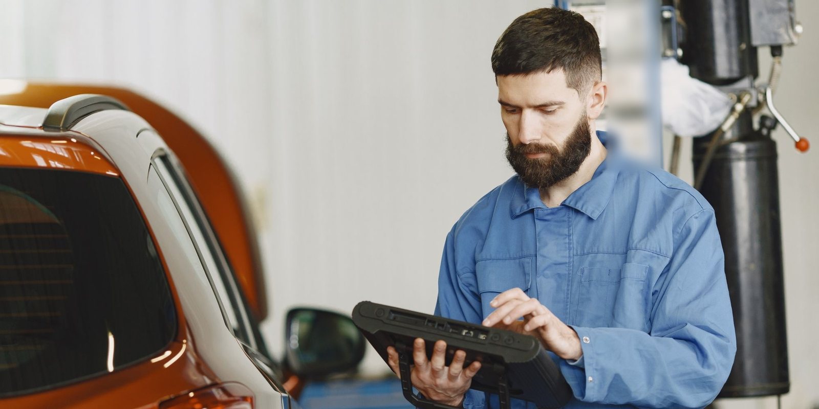 Técnico de uniforme azul consulta um tablet ao lado da traseira de um carro laranja dentro da oficina